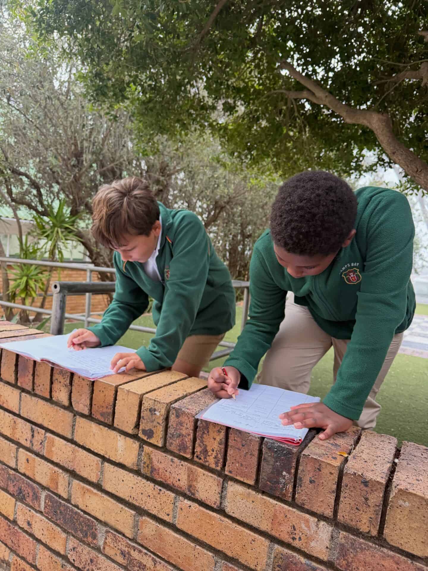 Two learners working on worksheets outdoors on a garden wall